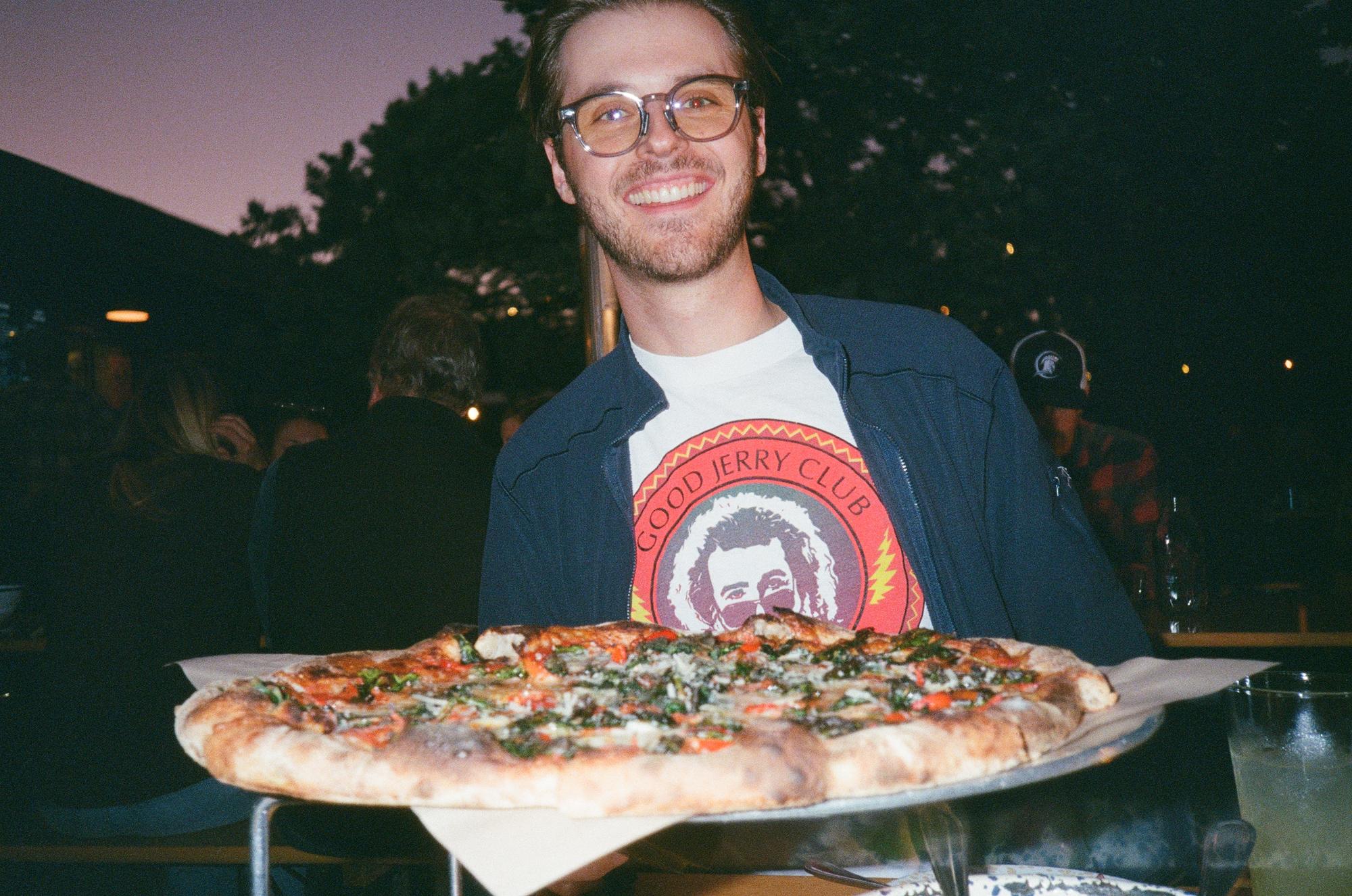 Jake Heller grinning behind an enormous pizza on a metal tray at an outdoor table in the evening